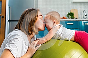 Close up portrait of a young mother leaning on a fit ball and kisses her baby. The concept of fitness with children at home