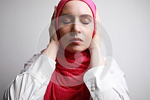 Close-up portrait of young Muslim woman in hijab closing her ears and eyes posing against wall. Space for text.