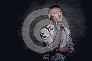 Close-up portrait of a young old-fashioned tattooed guy wearing white shirt and suspenders. Isolated on dark background.