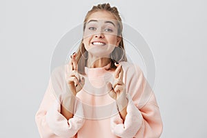 Close up portrait of young pretty superstitious woman in pink sweatshirt looking at the camera crossing her fingers for