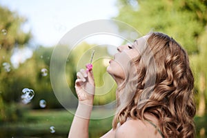 Close-up portrait of young pretty woman with blond curly hair, blowing soap bubbles in green park in summer. Summertime fun.