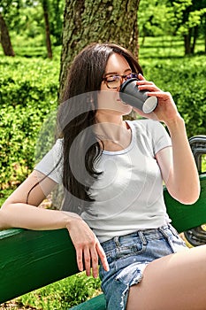 Close-up portrait young pretty woman drinking coffee from black paper cup sitting on a bench in the park