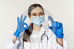 Close up portrait of young professional woman doctor, physician in medical face mask and glasses, showing vaccine and
