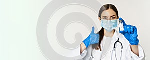 Close up portrait of young professional woman doctor, physician in medical face mask and glasses, showing vaccine and