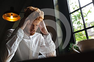 Close-up portrait of young readhead bearded overworked man in white shirt touching his head while sitting at office