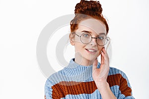 Close up portrait of young redhead woman in glasses touching her face and smiling. Girl with hairbun wearing eyewear and