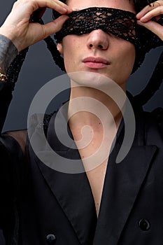 Close-up portrait of young sexy feminine male in black suit with eye mask, isolated on black