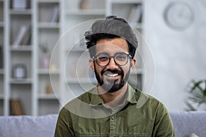 Close-up portrait of young smiling hispanic man smiling and looking at camera while sitting on sofa in living room