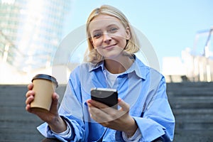 Close up portrait of young smiling woman with cup of coffee, drinking and sitting on stairs in city, holding smartphone