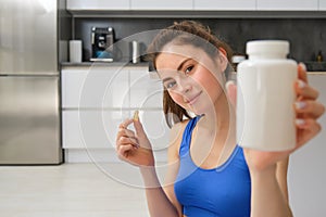 Close up portrait of young woman, fitness instructor showing bottle of vitamins, taking buds supplementary, dietary pill