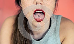 Close up portrait of young woman with freckles sticking out pierced tongue, showing her tongue piercing with pink background