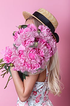 Close-up portrait of young woman in summer dress and straw hat holding peonies bouquet over her shoulder