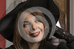 Close-up portrait of young woman wearing black wide-brimmed hat with veil, lace gloves, red lipstick. Girl from high society