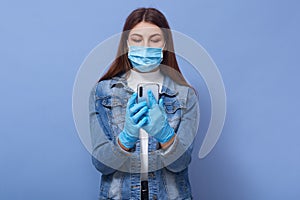 Close up portrait of young woman wearing medical face mask and blue gloves typing on mobile phone isolated over lilac background,