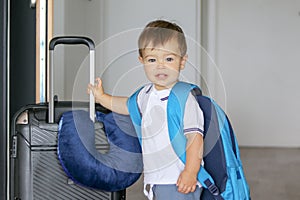 Close-up portrat of cute smiling little baby boy with big backpack and suitcase with travelling pillow stay near open door ready t
