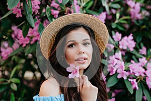 Close up potrait of beautiful young brunette woman in straw hat with pink flowers near her face