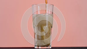 Close up of pouring cola in a long transparent glass with Ice and bubbles. Action. Soda soft drink isolated on pink wall