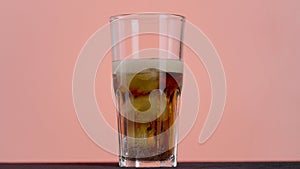 Close up of pouring cola in a long transparent glass with Ice and bubbles. Action. Soda soft drink isolated on pink wall