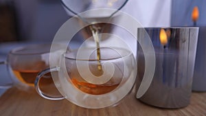 Close-up pouring healthful tea in transparent cup on table with burning candle. Unrecognizable Caucasian person