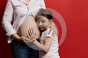 Close-up pregnant mother gently hugging her daughter who is touching bare belly,  on red background