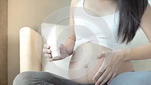 Close up of pregnant woman applying moisturizing cream on her belly  sitting on bed.