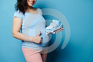 Close-up pregnant woman with big belly, shows thumb up, smiles at camera, holding newborn baby clothes, isolated on blue