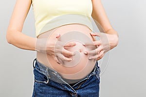 Close up of pregnant woman dressed in opened jeans scratching her belly at colorful background with copy space. Problem of stretch