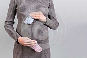 Close up of pregnant woman in gray dress holding baby pink and blue socks against her belly at gray background. Expecting twins.