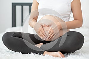 Close up pregnant woman hands touch her belly and make symbol of heart also sit on white bed with morning light. Concept of good