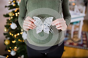 Close up of pregnant woman holding butterfly christmas ornament on belly against christmas tree in the background