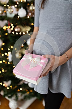 Close up of pregnant woman holding gift box against christmas tree in the background. Its a girl concept