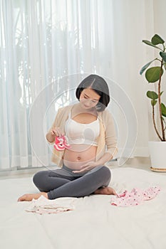 Close up of pregnant woman in home clothing holding shoes for a baby girl against her abdomen