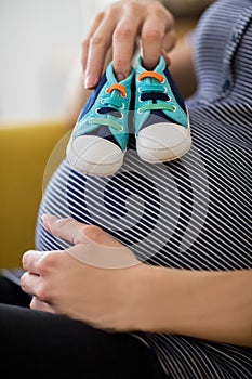 Close Up Of Pregnant Woman Sitting On Sofa At Home Holding Baby Shoes On Stomach
