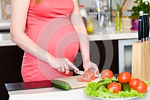 Close up pregnant woman with knife on kitchen cuts tomato