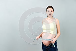 Close up of a pregnant woman in opened jeans measuring her belly with a tape measure at colorful background with copy space.