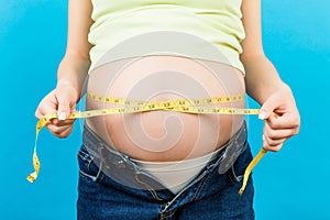 Close up of a pregnant woman in opened jeans measuring her belly with a tape measure at colorful background with copy space. Inch
