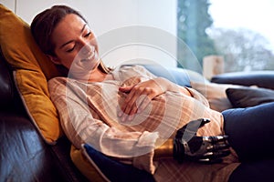 Close Up Of Pregnant Woman With Prosthetic Arm Relaxing Lying On Sofa At Home Holding Bump