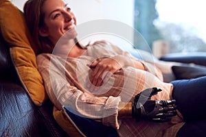 Close Up Of Pregnant Woman With Prosthetic Arm Relaxing Lying On Sofa At Home Holding Bump