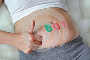 Close up Pregnant woman sitting on soft sofa and touching her belly with sign BOY in front of her belly. Showing thumbs up sign