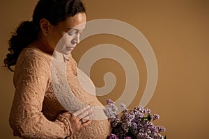 Close-up pregnant woman stroking her tummy, holding bouquet of purple blooming lilacs, isolated beige studio background