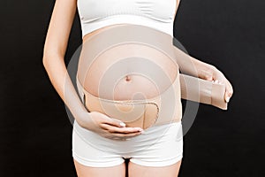 Close up of pregnant woman in underwear dressing pregnancy corset on the third trimester at black background with copy space.