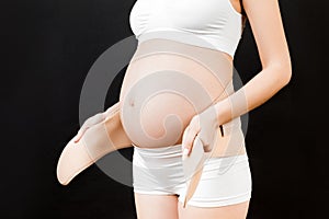 Close up of pregnant woman in underwear putting on pregnancy bandage at black background with copy space. Orthopedic abdominal