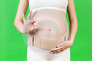 Close up of pregnant woman in underwear wearing supporting bandage and showing okay gesture at green background with copy space.