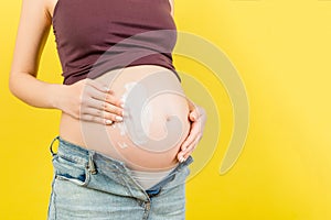 Close up of pregnant woman in unzipped jeans applying moisturizing cream on her belly against stretch marks at colorful background