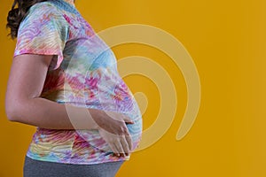Close up of pregnant woman wearing hipster tie dye t-shirt and grey yoga pants, arms on her belly. Female hands wrapped around big