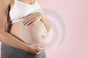 Close up of pregnant woman wearing supportive seamless maternity bra & grey yoga pants, arms on her belly. Female hands wrapped