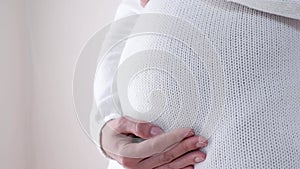 Close-up of a pregnant woman in a white sweater stroking her pregnant belly. Image of pregnancy and expectation of a