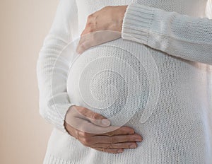 Close-up of a pregnant woman in a white sweater stroking her pregnant belly. Image of pregnancy and expectation of a