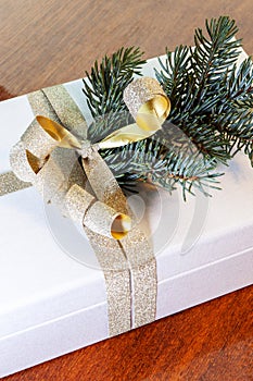 Close-up of present on a wooden vintage table. White gift box with golden bow and branch of Christmas tree.