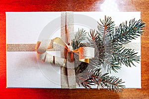 Close-up of present on a wooden vintage table. White gift box with golden bow and branch of Christmas tree.